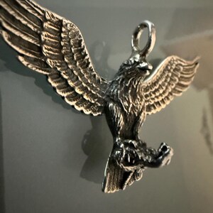 Może przedstawiać: Szczegółowa srebrna zawieszka w kształcie orła z rozłożonymi skrzydłami. Zawieszka posiada skomplikowane detale piór i pętlę na łańcuszek. Orzeł jest osadzony, z widocznymi szponami. Zawieszka jest prawdopodobnie wykonana ze srebra próby 925.