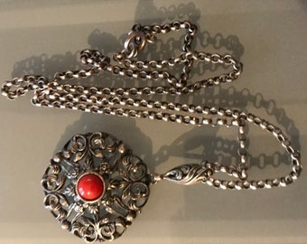 May include: Silver-toned necklace featuring a round pendant with intricate floral filigree and a central red stone. The chain is a delicate link style, and the pendant has a detailed, ornate design. The necklace is displayed on a neutral surface.