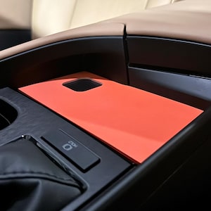 Puede incluir: Una placa de cubierta rectangular de color naranja brillante colocada dentro de la consola central de un vehículo. La cubierta presenta un pequeño recorte cuadrado y está situada junto a una zona de cambio de marchas negra con un botón etiquetado HOLD y tapicería de cuero beige.