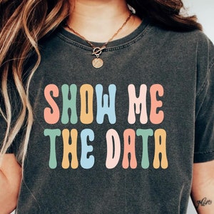 May include: A dark grey t-shirt with the text "SHOW ME THE DATA" in colorful, retro-style lettering. The shirt is made of a soft, comfortable fabric and has a relaxed fit.