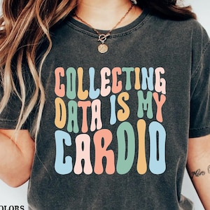 May include: A black t-shirt with the text "COLLECTING DATA IS MY CARDIO" in colorful, retro-style lettering. The shirt is worn by a person with long blonde hair and a necklace.