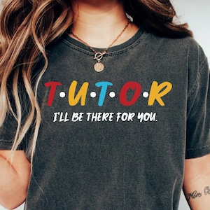 May include: A dark grey t-shirt with the text "T.U.T.O.R. I'll be there for you." in colorful letters. The shirt is made of a soft, comfortable material and has a relaxed fit.