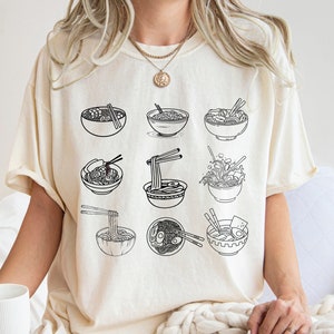 May include: A cream colored t-shirt with a black graphic of nine bowls of ramen noodles. The text "COMFORT COLORS IVORY" is at the bottom of the image.