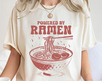 May include: A white t-shirt with the text "Powered by Ramen" in red. The shirt features a vintage-style illustration of a bowl of ramen noodles with chopsticks.