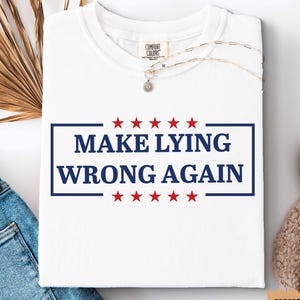 May include: White t-shirt with the text "MAKE LYING WRONG AGAIN" in a blue rectangle, with red stars above and below the text. The shirt is folded and placed on a white surface, with a pair of blue jeans and a white shoe visible.