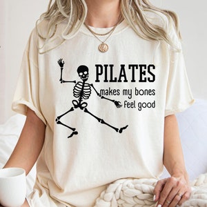 May include: Ivory-colored t-shirt with the text "PILATES makes my bones feel good" and a black skeleton graphic. The shirt is a Comfort Colors brand.
