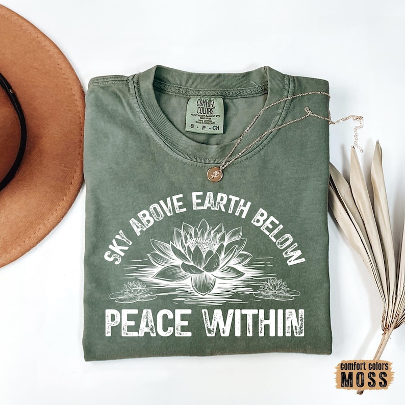 May include: A green t-shirt with the text "Sky Above Earth Below Peace Within" in white. A white lotus flower is in the center of the text. The shirt is made by Comfort Colors Moss.
