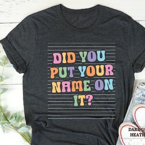 May include: Dark grey heather t-shirt with the text "Did you put your name on it?" in colorful letters.