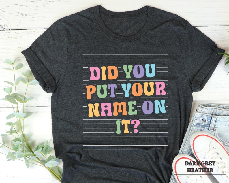 May include: Dark grey heather t-shirt with the text "Did you put your name on it?" in colorful letters.