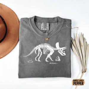 Może przedstawiać: Szary t-shirt z nadrukiem białego konturu szkieletu dinozaura. Dinozaur to Triceratops. Na metce t-shirtu widnieje napis "Comfort Colors".