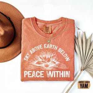 May include: A coral-colored t-shirt with a white graphic design that reads "Sky Above Earth Below Peace Within". The graphic features a lotus flower and smaller lotus flowers.