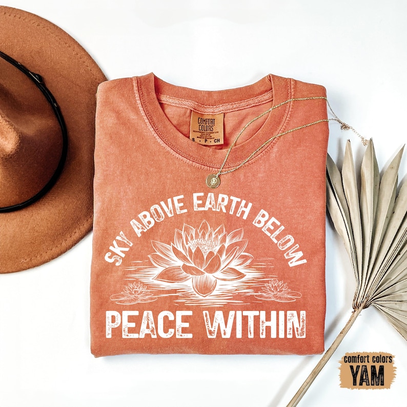 May include: A coral-colored t-shirt with a white graphic design that reads "Sky Above Earth Below Peace Within". The graphic features a lotus flower and smaller lotus flowers.