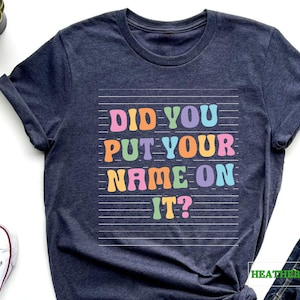 May include: Heather navy blue t-shirt with the text "Did you put your name on it?" in rainbow colors on a white lined background.