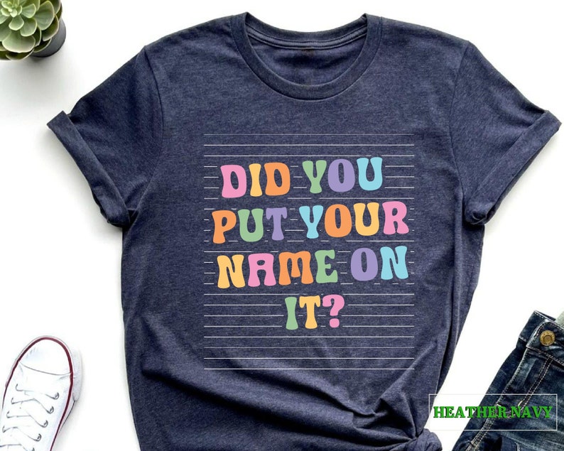 May include: Heather navy blue t-shirt with the text "Did you put your name on it?" in rainbow colors on a white lined background.