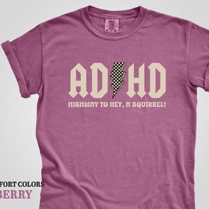 May include: A pink t-shirt with a white graphic that says "ADHD Highway to hey, a squirrel!" with a checkered lightning bolt.