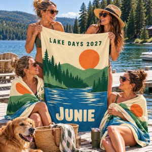 May include: A rectangular beach towel featuring a graphic print of a lake, pine trees, and a large orange sun. The text "LAKE DAYS 2027" is printed at the top, and the name "JUNIE" is printed in large, bold letters at the bottom of the towel.