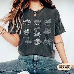 May include: A dark gray t-shirt with a white graphic of multiple bowls of ramen noodles. The shirt has a distressed look and is made of a soft, comfortable fabric. The text "COMFORT COLORS PEPPER" is visible in the lower right corner of the image.