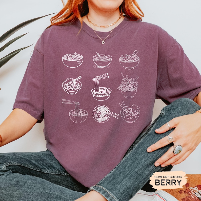 May include: A purple t-shirt with a white graphic of nine bowls of ramen noodles. The graphic is centered on the shirt. The shirt has the words "Comfort Colors Berry" printed on the bottom right corner.