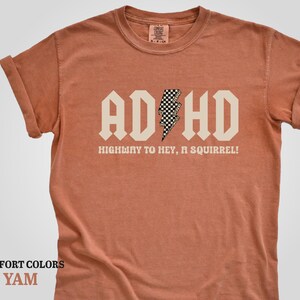 May include: A rust-colored t-shirt with a white graphic that says "ADHD" in a bold font with a checkered lightning bolt in the middle. The text below the graphic says "Highway to hey, a squirrel!"