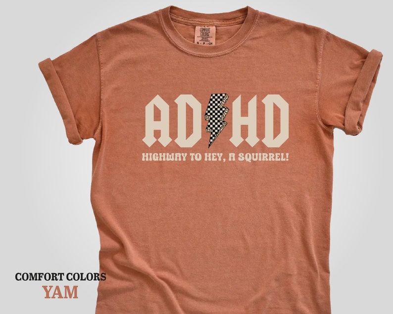 May include: A rust-colored t-shirt with a white graphic that says "ADHD" in a bold font with a checkered lightning bolt in the middle. The text below the graphic says "Highway to hey, a squirrel!"