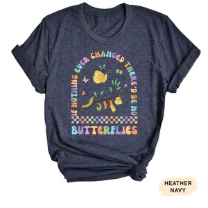 K&ouml;nnte beinhalten: Ein heidefarbenes marineblaues T-Shirt mit einer bunten Grafik eines Zweigs mit Schmetterlingen und Raupen. Der Text auf dem Shirt lautet "If nothing ever changed there'd be no butterflies".