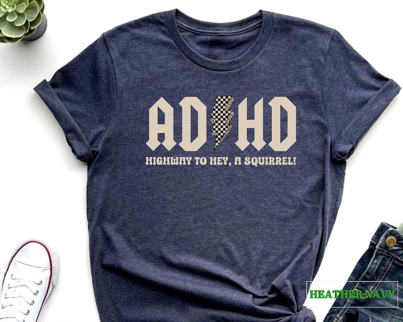 May include: Heather navy blue t-shirt with a beige graphic that says "ADHD Highway to Hey, a Squirrel!" with a black and white checkered lightning bolt.