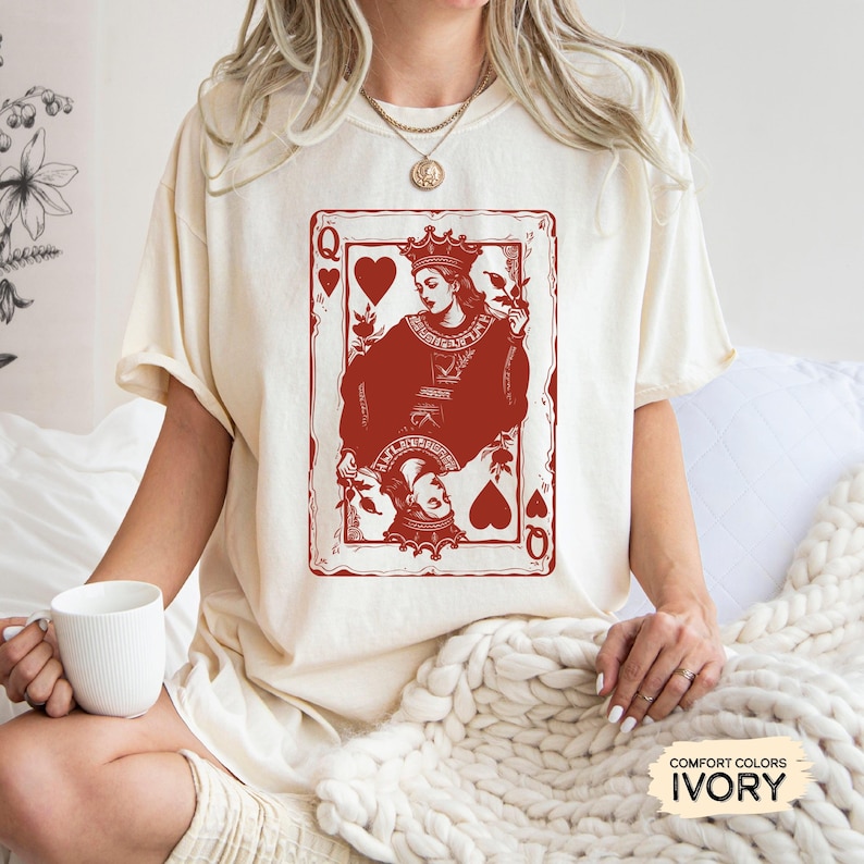 May include: A white t-shirt with a red Queen of Hearts playing card graphic. The card features a stylized illustration of a queen holding a flower. The text "COMFORT COLORS IVORY" is visible in the bottom right corner of the image.