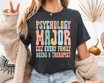 May include: Dark grey heather t-shirt with the text "PSYCHOLOGY MAJOR CUZ EVERY FAMILY NEEDS A THERAPIST" in colorful, retro-style lettering. The shirt is a crew neck and has short sleeves.