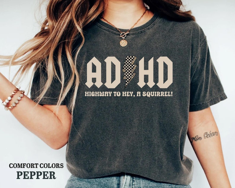 May include: A dark gray t-shirt with a beige graphic that says "ADHD Highway to Hey, a Squirrel!" The graphic features a black and white checkered lightning bolt.