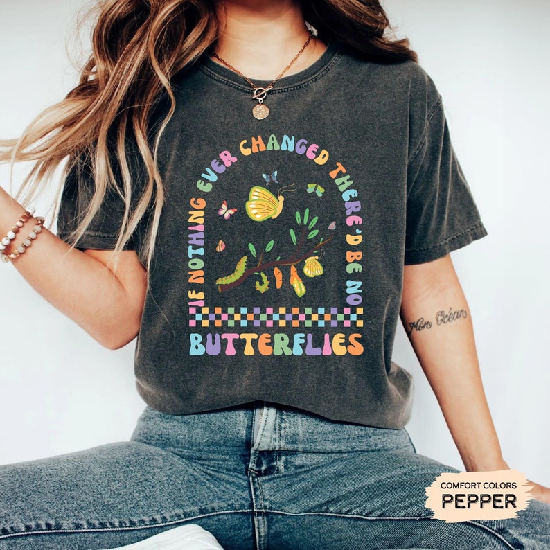 K&ouml;nnte beinhalten: Ein dunkelgraues T-Shirt mit einer bunten Grafik, auf der "Wenn sich nichts jemals &auml;ndern w&uuml;rde, g&auml;be es keine Schmetterlinge" steht. Die Grafik zeigt einen Zweig mit einer Raupe, Schmetterlingen und Kokons.