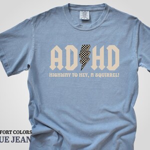 May include: A light blue t-shirt with a white graphic that says "ADHD" in a bold font with a checkered lightning bolt in the middle. The text below the graphic says "Highway to hey, a squirrel!"