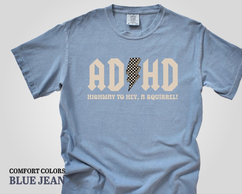 May include: A light blue t-shirt with a white graphic that says "ADHD" in a bold font with a checkered lightning bolt in the middle. The text below the graphic says "Highway to hey, a squirrel!"