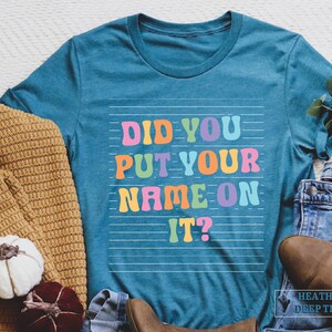 May include: A teal blue t-shirt with the text "Did you put your name on it?" in rainbow colors. The text is on a white background with thin white lines.
