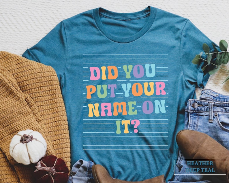 May include: A teal blue t-shirt with the text "Did you put your name on it?" in rainbow colors. The text is on a white background with thin white lines.