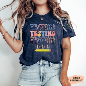 May include: Heather midnight navy t-shirt with the word "TESTING" in a retro font repeated three times in different colors. Below the word "TESTING" are the numbers 1, 2, and 3, and a yellow pencil.