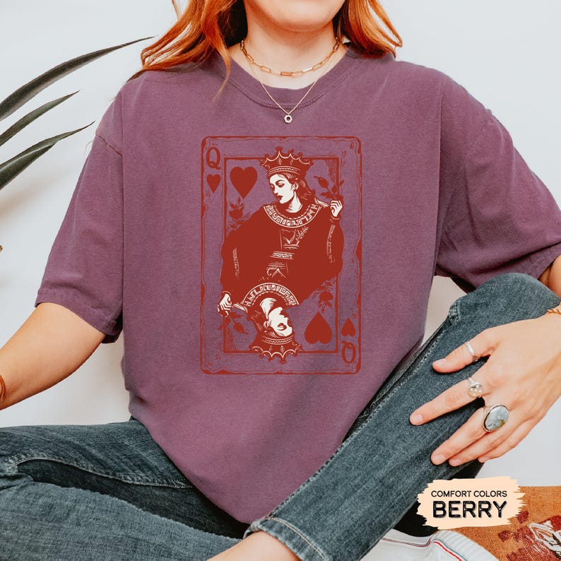 May include: A maroon t-shirt with a graphic of a queen of hearts playing card. The queen is depicted with a crown and holding a sword. The card is surrounded by a red border.