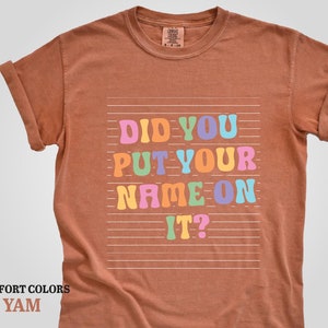 May include: A brown t-shirt with the text "Did you put your name on it?" in colorful letters. The text is on a white background with horizontal lines.