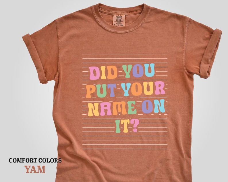 May include: A brown t-shirt with the text "Did you put your name on it?" in colorful letters. The text is on a white background with horizontal lines.
