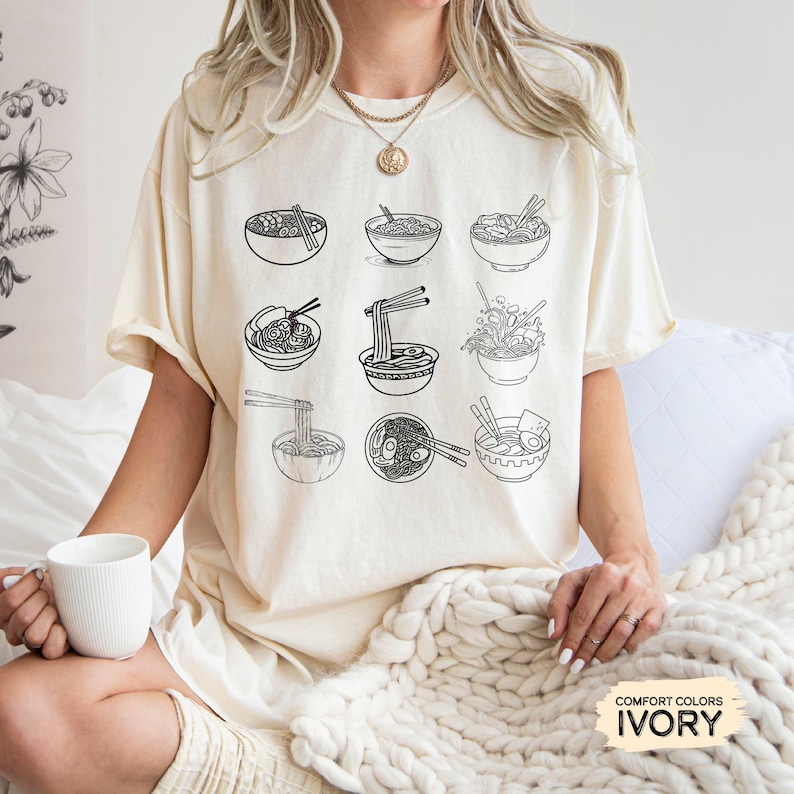 May include: A cream colored t-shirt with a black graphic of nine bowls of ramen noodles. The text "COMFORT COLORS IVORY" is at the bottom of the image.