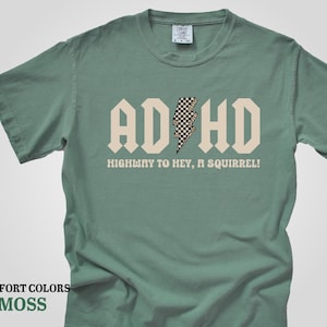May include: A moss green t-shirt with a white graphic that says "ADHD" with a checkered lightning bolt in the middle. The text below the graphic says "Highway to hey, a squirrel!"