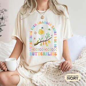 K&ouml;nnte beinhalten: Ein wei&szlig;es T-Shirt mit einer bunten Grafik, auf der steht: "Wenn sich nichts jemals &auml;ndern w&uuml;rde, g&auml;be es keine Schmetterlinge". Die Grafik zeigt Schmetterlinge, Raupen und Kokons.