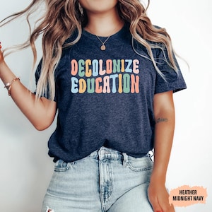 May include: Heather midnight navy t-shirt with the text "DECOLONIZE EDUCATION" in colorful, rounded letters. The shirt is a dark blue color and is paired with light blue denim jeans.
