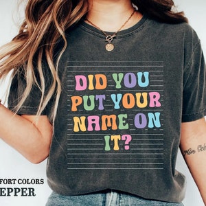 May include: A dark gray t-shirt with a colorful graphic that says "Did you put your name on it?" The graphic is on a white background with black lines.