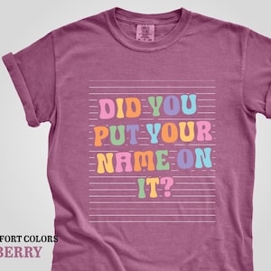 May include: A berry colored t-shirt with the text "Did you put your name on it?" in rainbow colors on a white background.