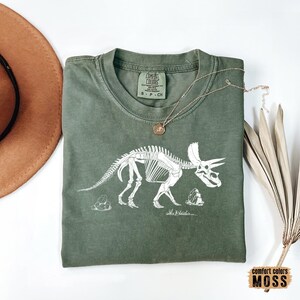 Może przedstawiać: Zielona koszulka z białym obrysem szkieletu dinozaura. Dinozaur to Triceratops. Koszulka ma napis "Comfort Colors" na metce.