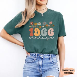 May include: Teal-colored t-shirt with a floral design above the text "1966 vintage". The numbers are in orange, yellow, and pink. The word "vintage" is in a cursive font. The shirt is paired with blue denim shorts.