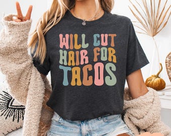 May include: Dark grey heather t-shirt with the text "WILL CUT HAIR FOR TACOS" in colorful retro font. The shirt is a crew neck and has short sleeves. The text is in a groovy, retro style.