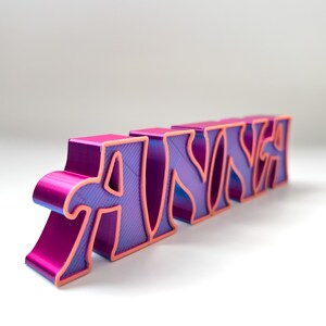 K&ouml;nnte beinhalten: Ein 3D-gedrucktes Namensschild mit der Aufschrift "ANWYA" in Pink und Lila mit pinkfarbenem Umriss. Die Buchstaben sind stilisiert und haben eine strukturierte Oberfl&auml;che.