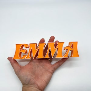 K&ouml;nnte beinhalten: Orangefarbene und wei&szlig;e 3D-Buchstaben, die den Namen "EMMA" buchstabieren.