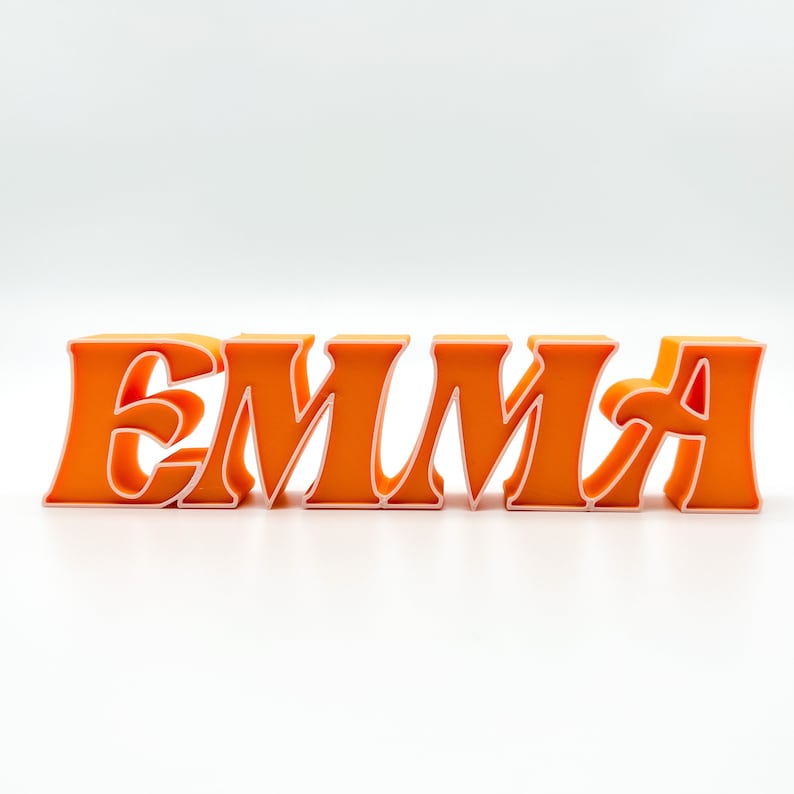K&ouml;nnte beinhalten: Orangefarbenes 3D-Namensschild mit der Aufschrift "EMMA" mit wei&szlig;em Umriss.